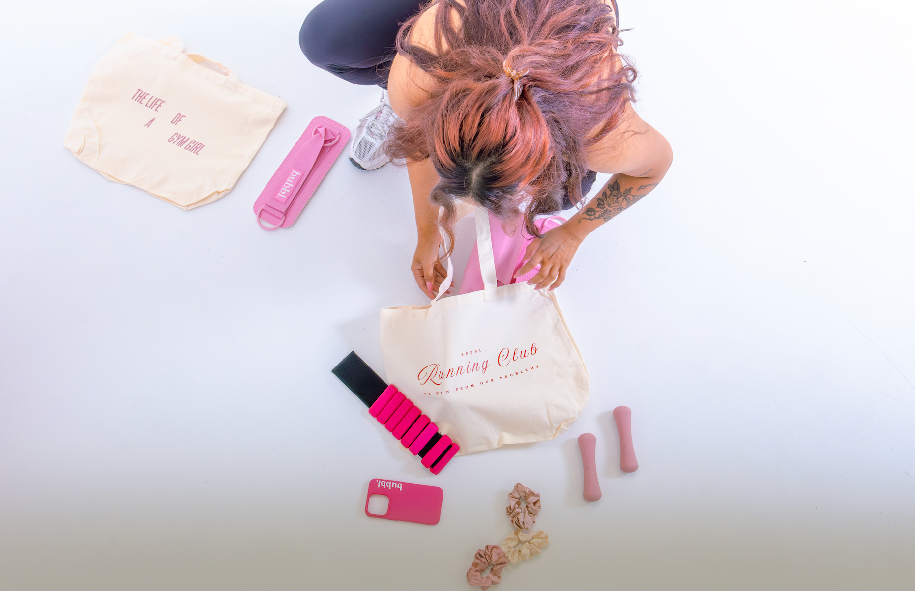 Person PACKING a tote bag with 'running club' branding, surrounded by pink gym accessories on a white background.